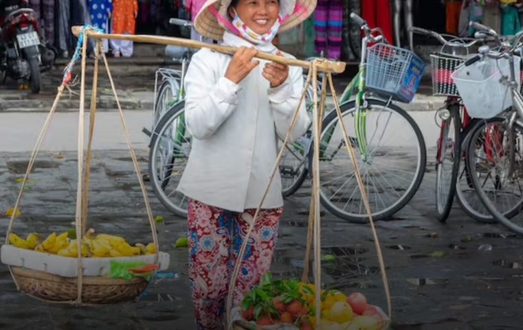A smiling woman in Vietnam with a traditional hat holds fruit and vegetables.