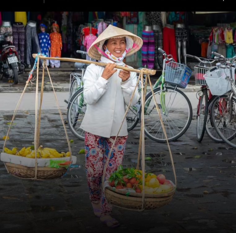 A smiling woman in Vietnam with a traditional hat holds fruit and vegetables.